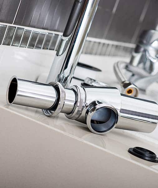 Siphon chrome pour lavabo : kit d'installation plomberie Composants de plomberie neufs en chrome brillant (siphon, raccords) posés sur un lavabo beige. Kit d'installation et réparation du drain.