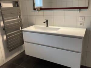 Meuble vasque blanc suspendu avec robinet noir, sèche-serviette gris et carrelage mural dans une salle de bain moderne.