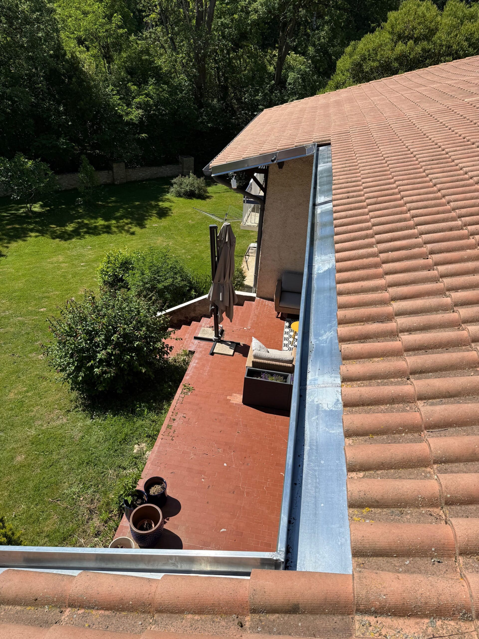 Gouttière en zinc sur toit de tuiles surplombant une terrasse et un jardin arboré.