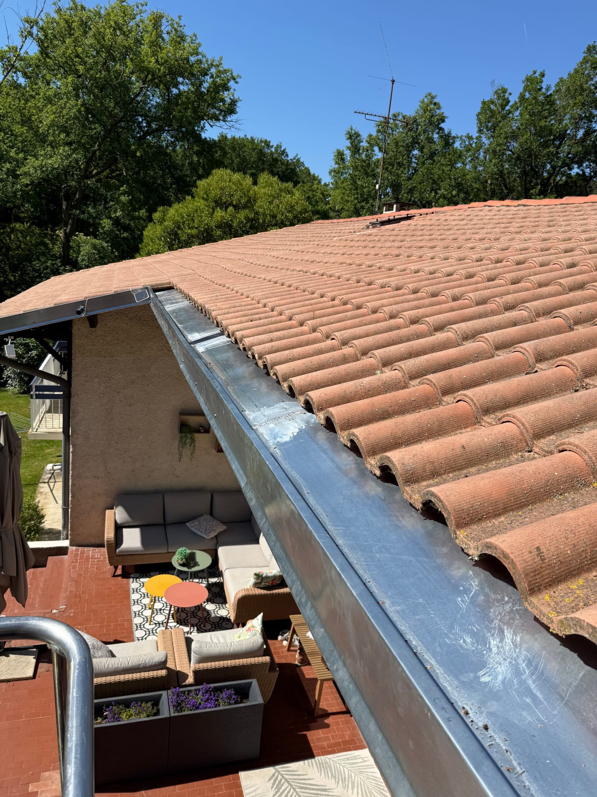 Toit de tuiles rouges et gouttière zinc dominant une terrasse avec mobilier de jardin moderne.
